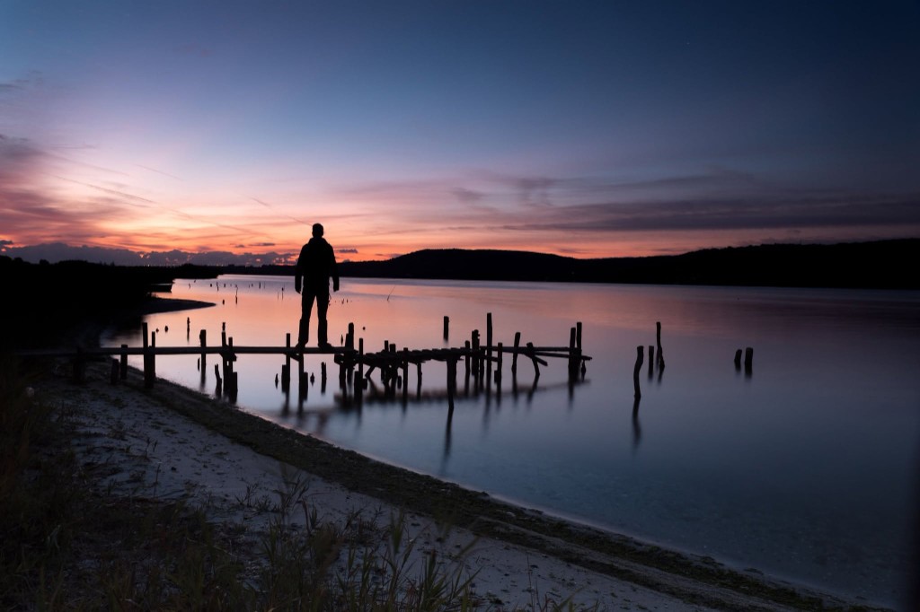 Silhouette of Todor Stoyanov standing on a wooden pier over calm water at sunset